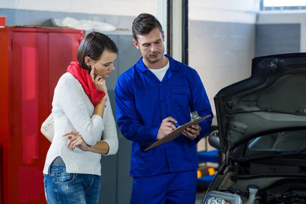 Auto mechanic explaining car repair details to a customer beside an open car hood in a service garage