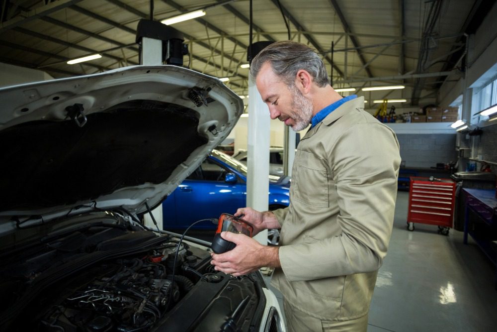 Mechanic in beige uniform inspecting a car engine with diagnostic tool in an auto repair workshop.