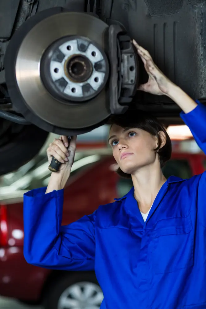 Female mechanic in blue uniform inspecting and repairing a car disc brake system in workshop.