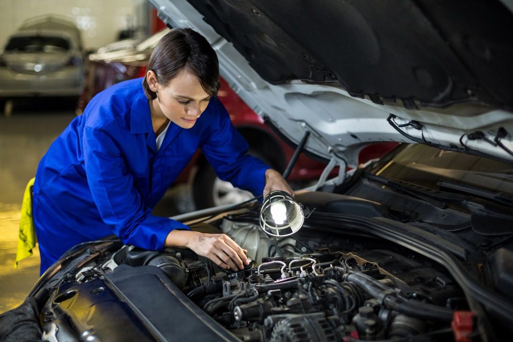 Mechanic in blue uniform inspecting car engine with flashlight in workshop for repair and service.