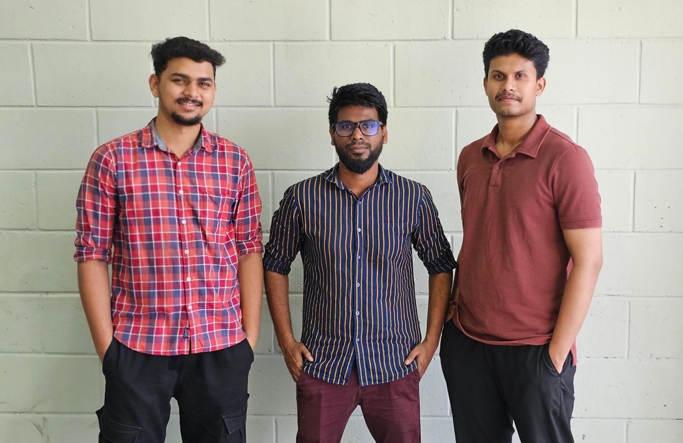 Three young men standing against a light brick wall, posing casually for a group photo