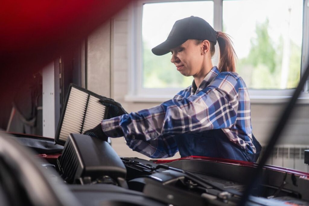 Female mechanic in cap and gloves replacing a car air filter during vehicle maintenance service.