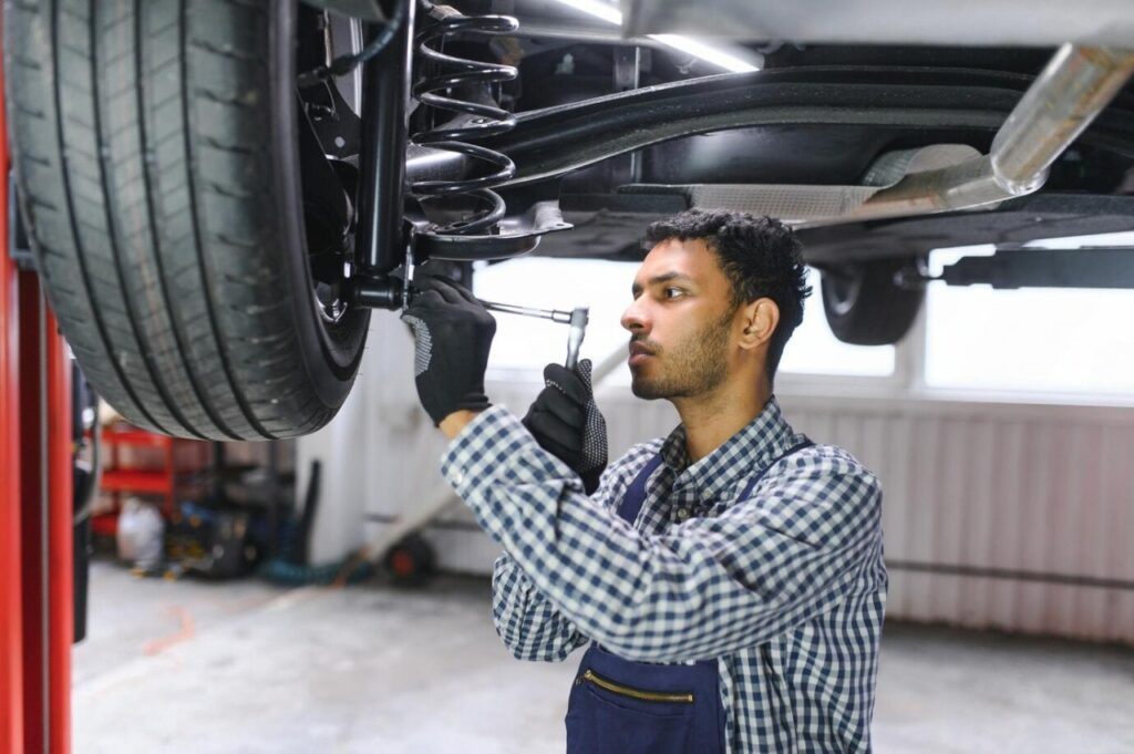 Mechanic in checkered shirt and gloves repairing car suspension under lifted vehicle in workshop.